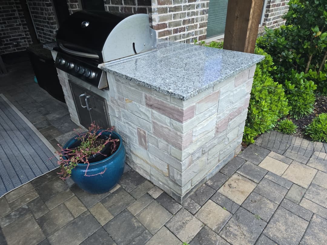 Outdoor kitchen with stone grill island and granite countertop beside a potted plant.