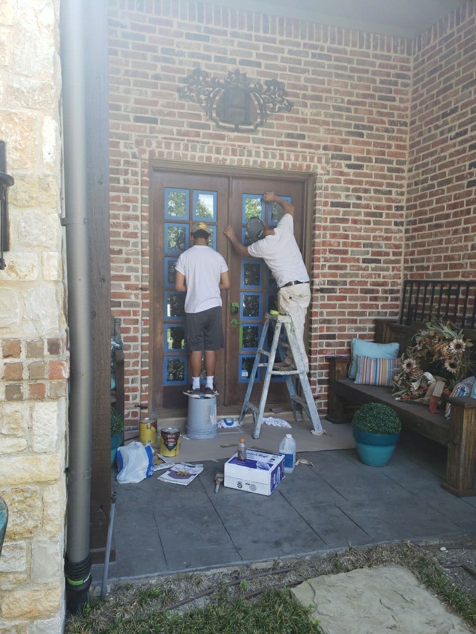 Two painters working on a decorative wooden front door, surrounded by tools and planters.