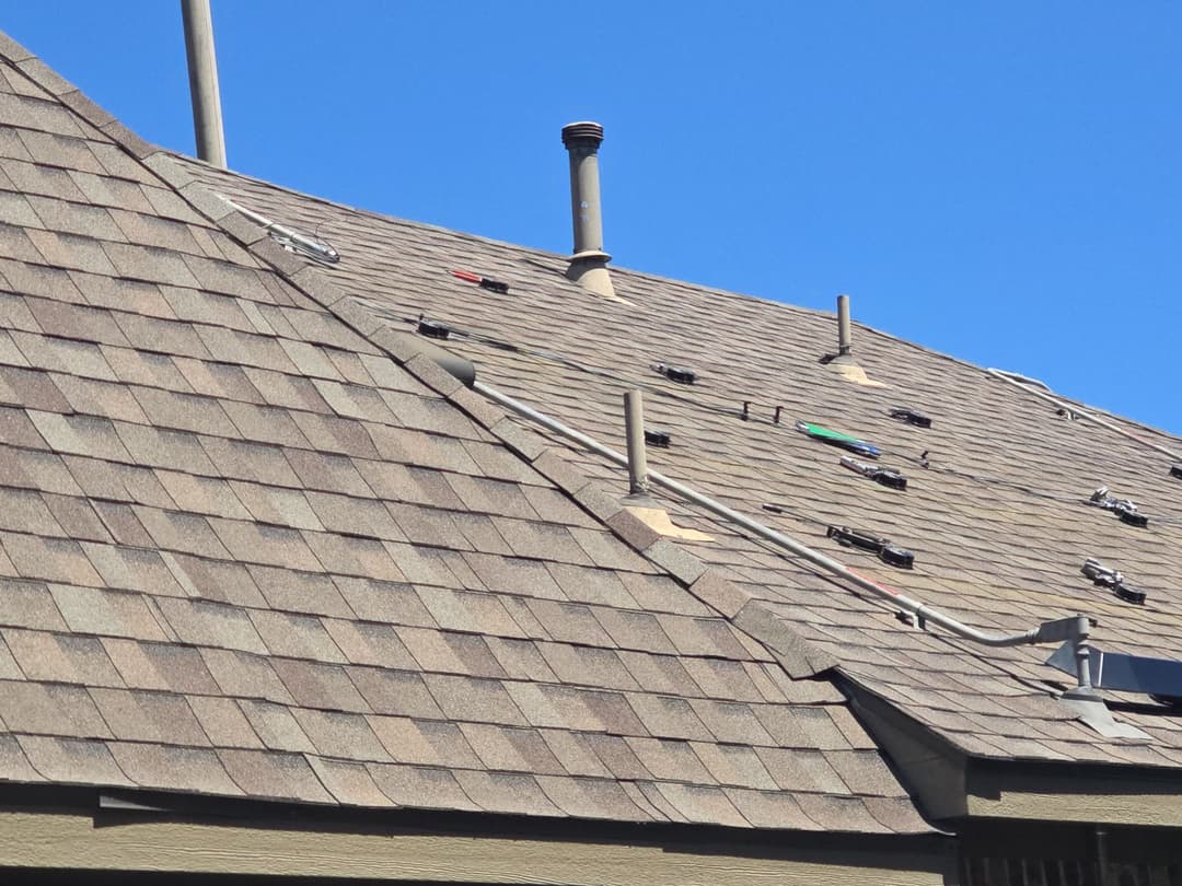 Roof shingles with ventilation pipes and tools against a clear blue sky.