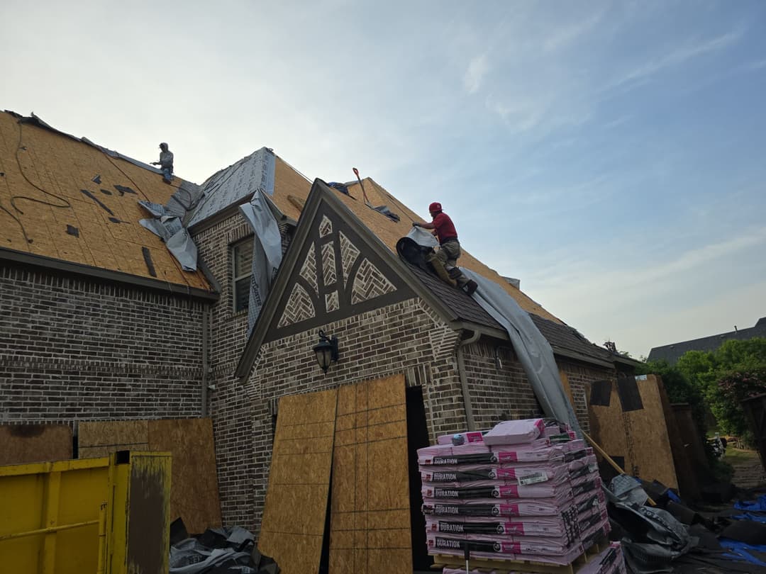 Roofers installing shingles on a house, with materials stacked nearby and construction visible.