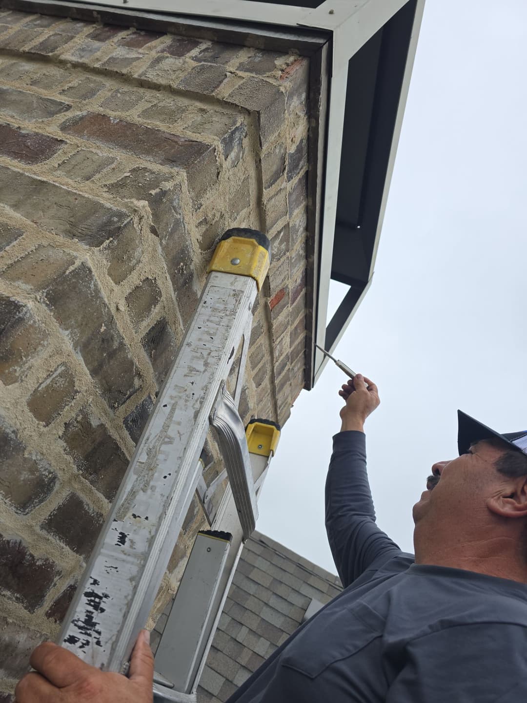 Man on ladder applying sealant to brick exterior of a house under overhang.