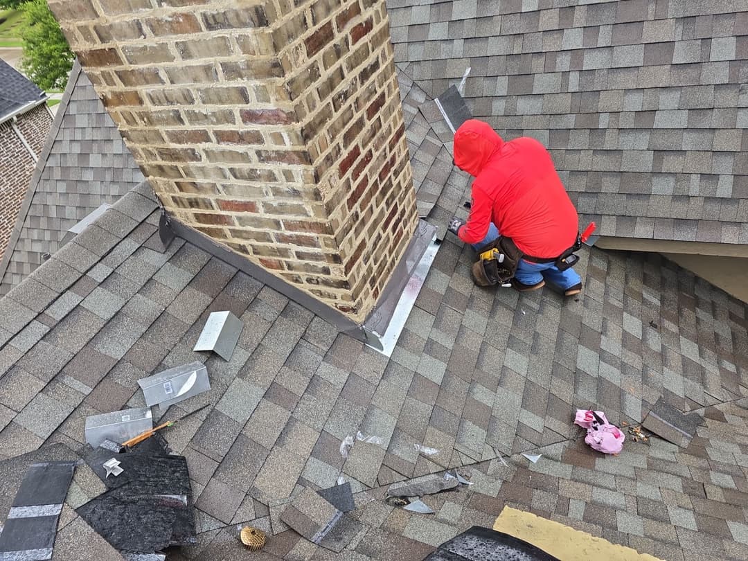 Roofer in red jacket repairing chimney on a shingled roof with construction tools nearby.