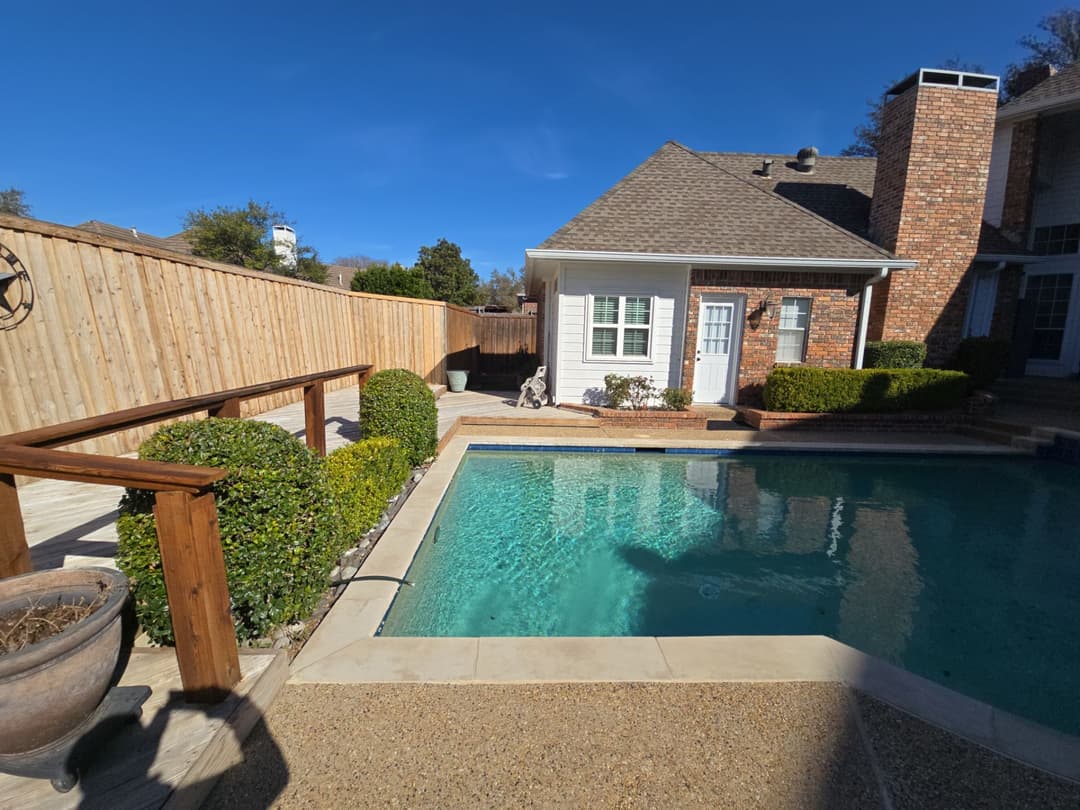 Backyard with a crystal clear pool, bordered by manicured hedges and a brick house in the background.