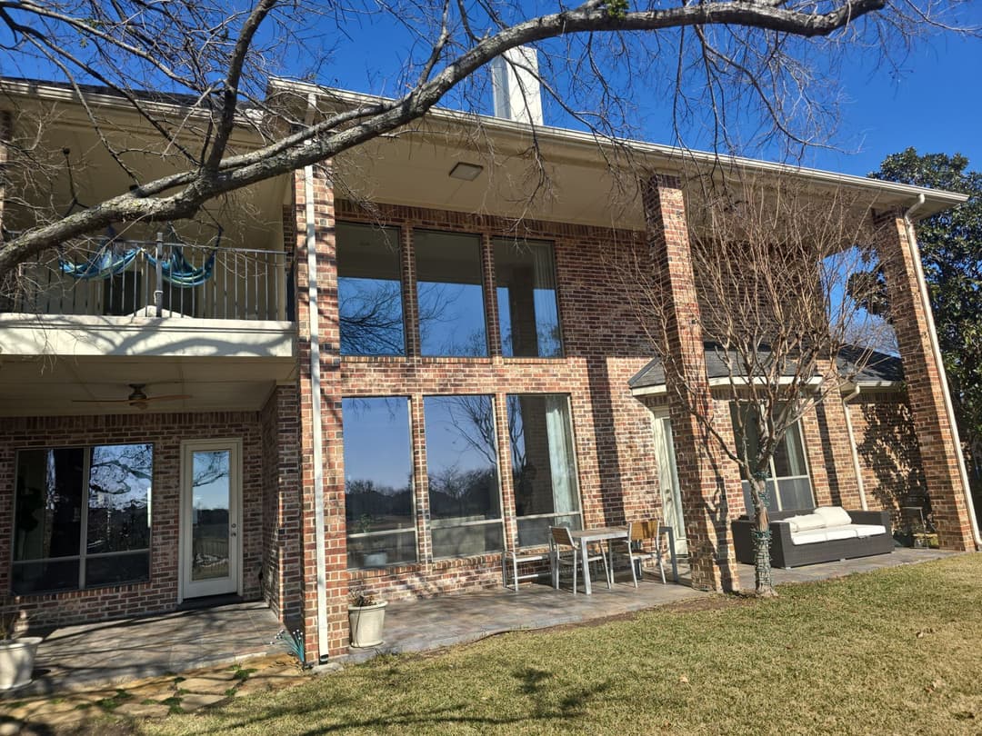 Modern brick house with large windows, patio seating, and leafy trees on a sunny day.