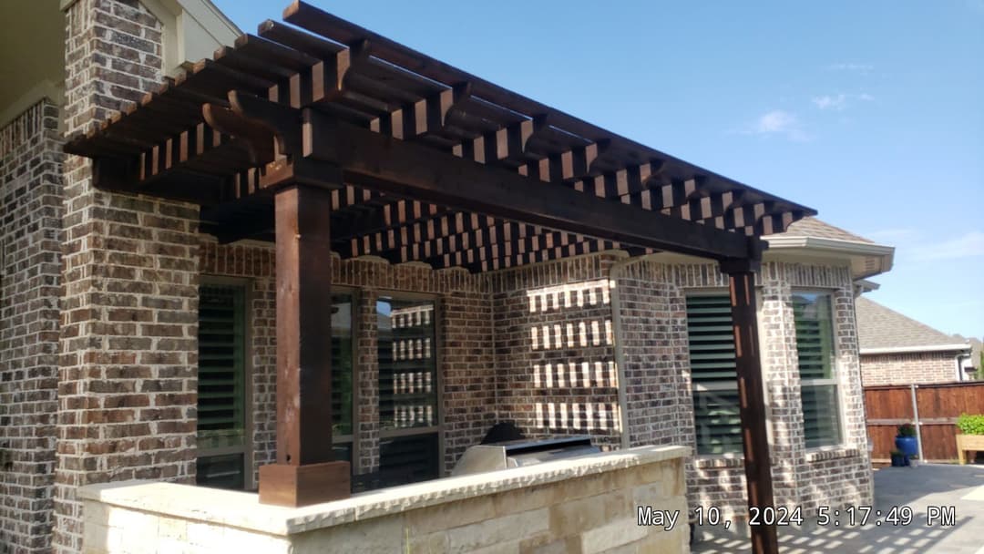 Wooden pergola attached to a brick house with outdoor seating area and clear blue sky.