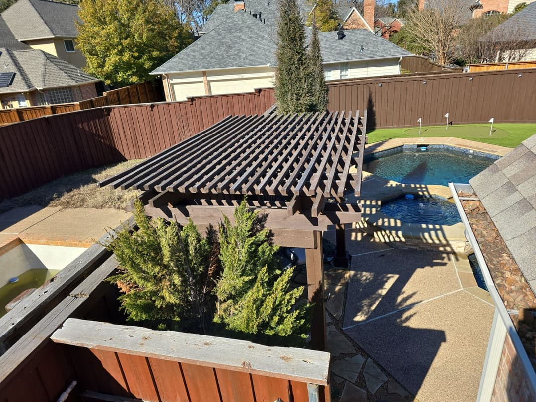Wooden pergola over patio by a pool in a landscaped backyard with shrubs and trees.