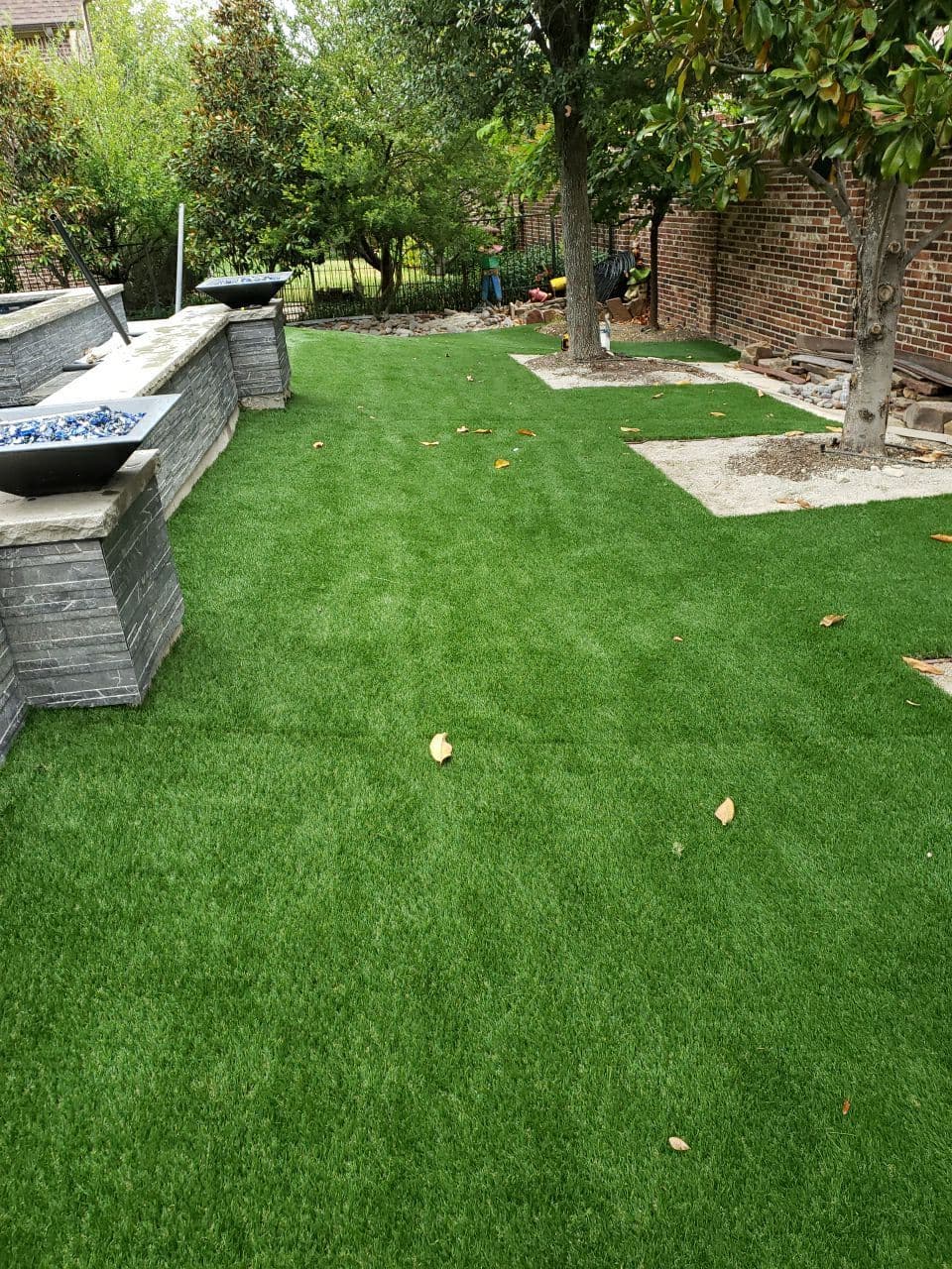 Lush green artificial grass in landscaped backyard with trees and stone features.